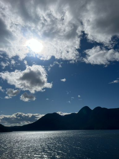 Helle Sonne über Bergen und einem glitzernden Gewässer, mit Wolken am Himmel.