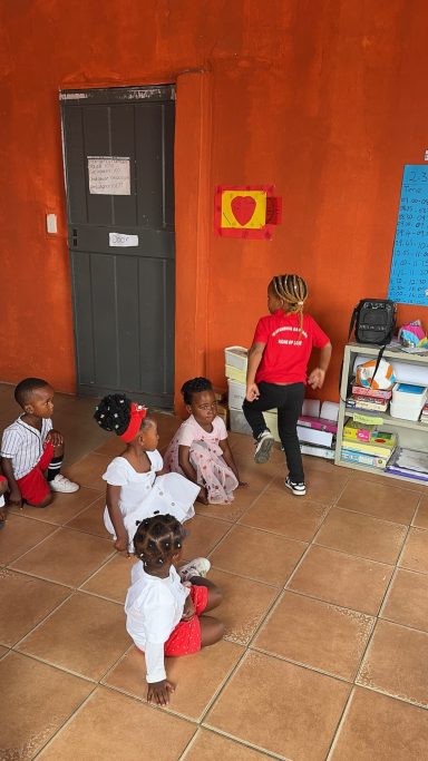 Gruppe von Kindern in einem Raum, einige sitzen, andere stehen, bunte Kleidung.