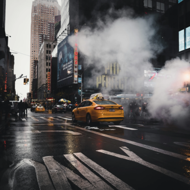 Gelbes Taxi fährt durch eine regnerische Stadtstraße mit dampfendem Asphalt.