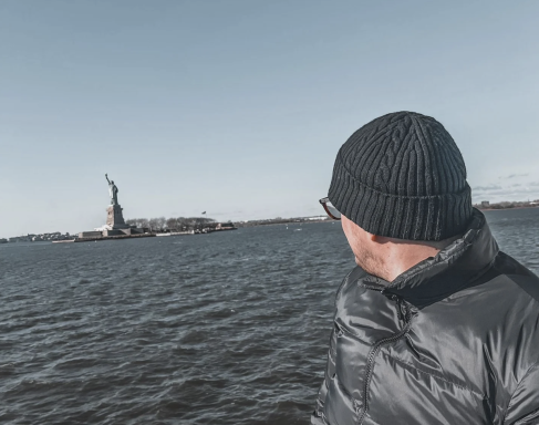 Person mit Mütze blickt auf die Freiheitsstatue vor ihm, umgeben von Wasser.
