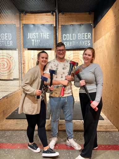 Drei Personen stehen in einer Wurf-Arena mit Äxten, lächelnd und bereit zum Werfen.