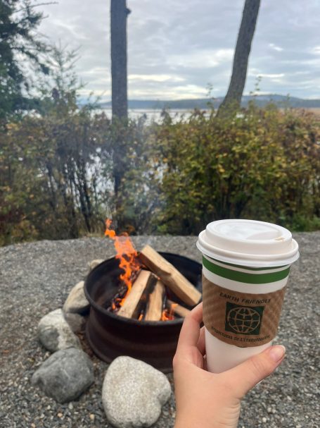 Eine Hand hält einen Kaffeebecher vor einem Lagerfeuer und einem See.