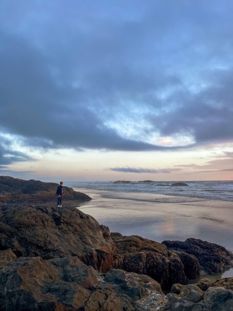 Person steht auf Felsen am Strand, Blick auf das Meer und bewölkten Himmel.